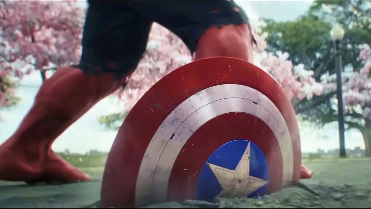 Captain America's shield is embedded in the pavement after a big impact. standing behind it, we see the legs of the Red Hulk